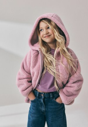 Młoda dziewczyna o długich blond włosach, ubrana w różową bluzę polarową z kapturem, fioletowy top i niebieskie dżinsy, ręce w kieszeniach, uśmiechająca się i patrząca na bok.
