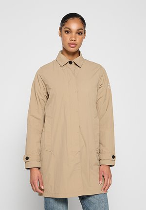 Kvinde iført en beige langærmet knapjakke over jeans, stående mod en ensfarvet lys grå baggrund.