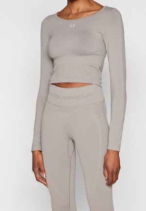 Femme portant un crop top ajusté à manches longues gris clair et un legging taille haute, tous deux de la marque Under Armour, sur un fond blanc uni.