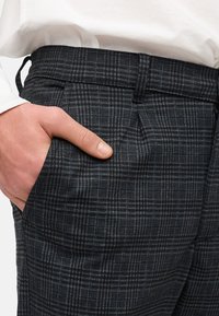 Wollen broek met een donker ruitpatroon. Heeft een vlakke voorkant, zijzakken en een klassieke pasvorm. Hand in de zak toont detail.