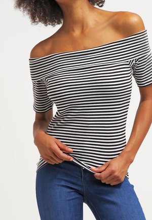 Vrouw met een zwart-wit gestreept off-shoulder topje en blauwe spijkerbroek, die met beide handen de zoom van het shirt aanpast.