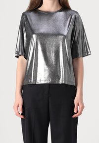 Top corto manica corta in argento metallico con scollatura rotonda e vestibilità rilassata. Il tessuto ha una finitura lucida, in contrasto con pantaloni neri.