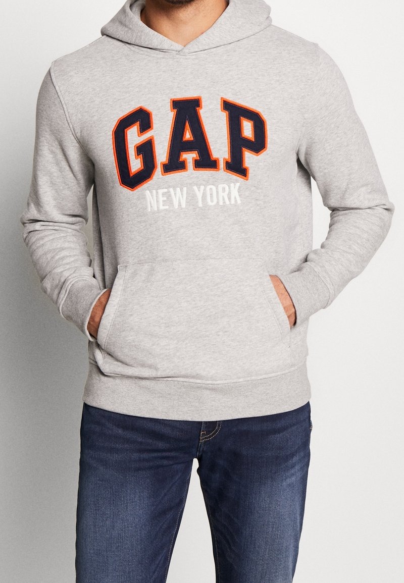 Homme portant un sweat à capuche gris clair GAP avec des lettres orange et bleu marine et le texte "New York", associé à un jean bleu foncé, mains dans les poches du sweat.