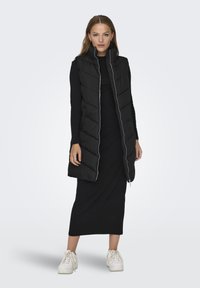 Gilet rembourré noir avec un design en losanges, col montant, fermeture éclair totale à l'avant et poches latérales, porté sur une longue robe noire et des baskets blanches.