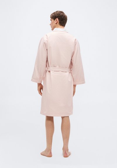 Άνδρας όρθιος, ξυπόλητος, φοράει ρόμπα μήκους έως τα γόνατα σε ανοιχτό ροζ χρώμα με ζώνη, απεικονιζόμενος από πίσω σε απλό λευκό φόντο.