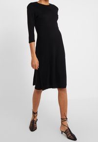 Robe noire de longueur genou avec des manches trois quarts, fabriquée dans un tissu doux. Dotée d'une fente sur le côté pour faciliter les mouvements. Assortie à des talons noirs à lanières.