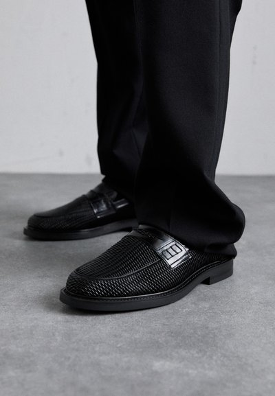 Schwarze, strukturierte Slipper mit einem glatten schwarzen Akzentband und einer flachen Sohle, kombiniert mit schwarzen Hosen, vor einem grauen Hintergrund.
