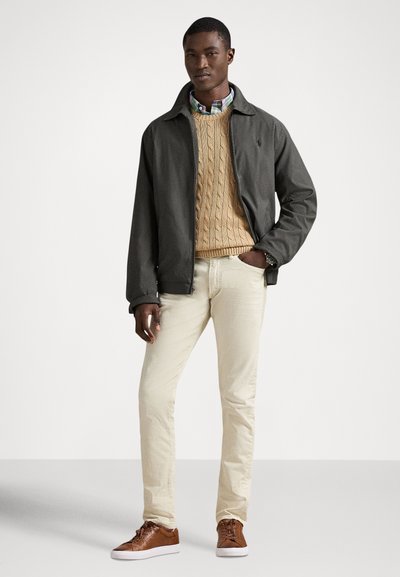 Chaqueta ligera gris oscuro sobre un suéter beige de punto trenzado, pantalones color crema y zapatillas marrones. Se ve el cuello de una camisa a cuadros multicolor.