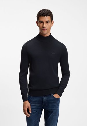 Mann mit lockigem Haar, der einen schwarzen Rollkragenpullover und blaue Jeans trägt, steht mit einer Hand in der Tasche vor einem schlichten weißen Hintergrund.