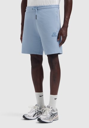Mann trägt hellblaue Shorts mit Kordelzug, weiße Nike-Socken und silberne Laufschuhe, steht vor einfachem Hintergrund.