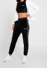 Černý sametový crop top a odpovídající teplákové kalhoty s nastavitelným pasem, zdobené logy a vzorovaným bočním pruhem. Bělé tenisky dotvářejí vzhled.