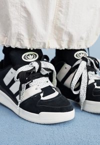 Black suede sneakers with white accents, featuring thick, textured laces and a distinctive logo on the tongue. Set against a soft blue background.