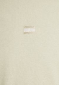Light green short-sleeve top made of soft fabric, featuring a square embroidered patch with beige and white stripes.
