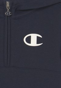 Mörkblå hoodie med en vit Champion-logotyp tryckt på bröstet och en dragkedja framtill med en metall Champion-logotypdragare.