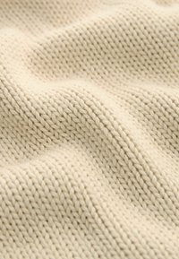Beige stickad tyg med en texturerad mönster, som visar mjuka, släta fibrer och en enhetlig, vågig yta. Inga synliga beslag eller accenter.