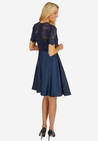 Navyblaues Kleid mit einem Spitzenoberteil mit gewelltem Rand, ausgestelltem Satinrock und kurzen Ärmeln; getragen mit schwarzen High Heels.