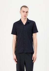 Camicia button-up a maniche corte di colore blu navy con tessuto testurizzato, dotata di colletto e taschino sul petto, abbinata a pantaloni a righe.