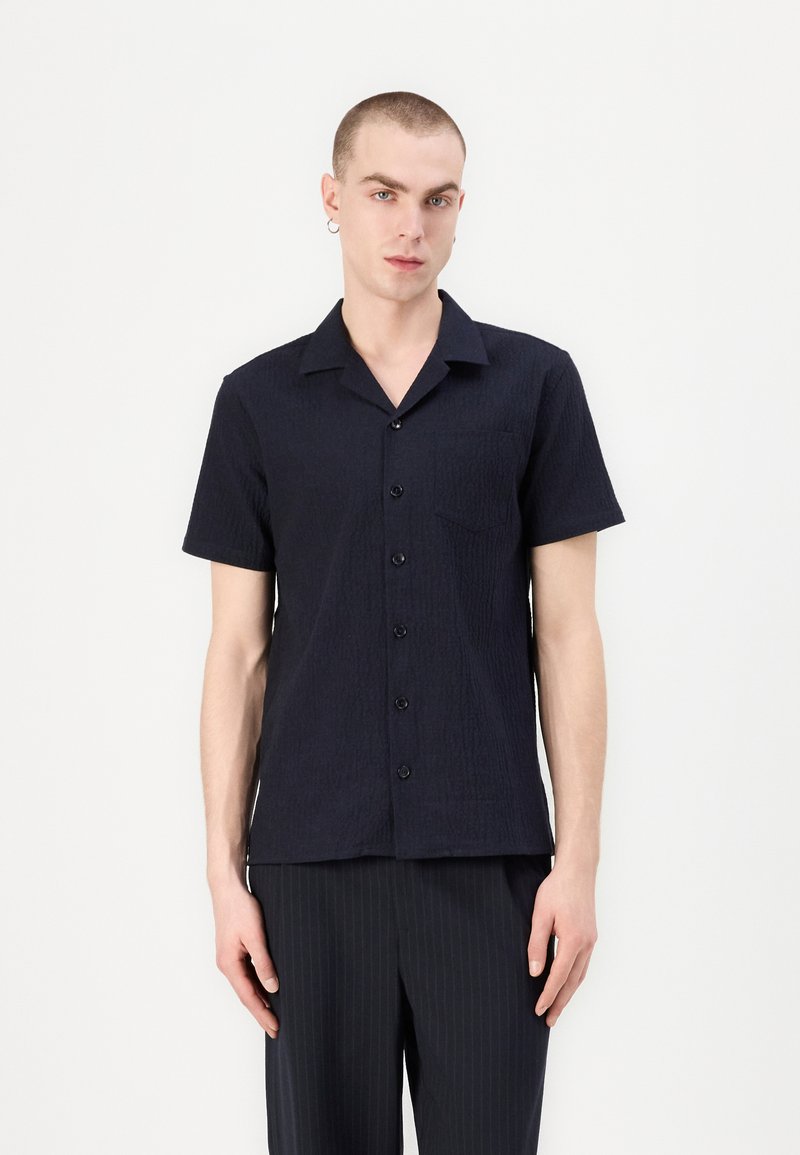 Camicia button-up a maniche corte di colore blu navy con tessuto testurizzato, dotata di colletto e taschino sul petto, abbinata a pantaloni a righe.