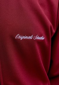 Μπορντό υφασμάτινη υφή με λευκό κεντημένο κείμενο με καλλιγραφική γραφή που λέει "Originals Studio" στην πάνω περιοχή ενός ενδύματος.