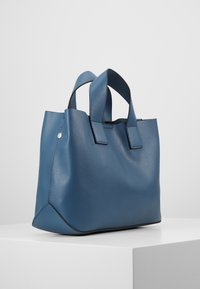 Modrá texturovaná kožená kabelka s dvojitými horními uchy, úhlovým tvarem a kontrastním černým lemem podél okrajů.