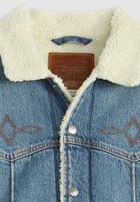 Denim jacket with a cream faux shearling collar. Features brown stitching, a leather label, and decorative diamond embroidery on the chest.