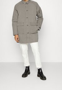 Abrigo de patrón a cuadros con dos bolsillos frontales, cierre de botones y cuello con solapas; combinado con pantalones blancos y botines negros.