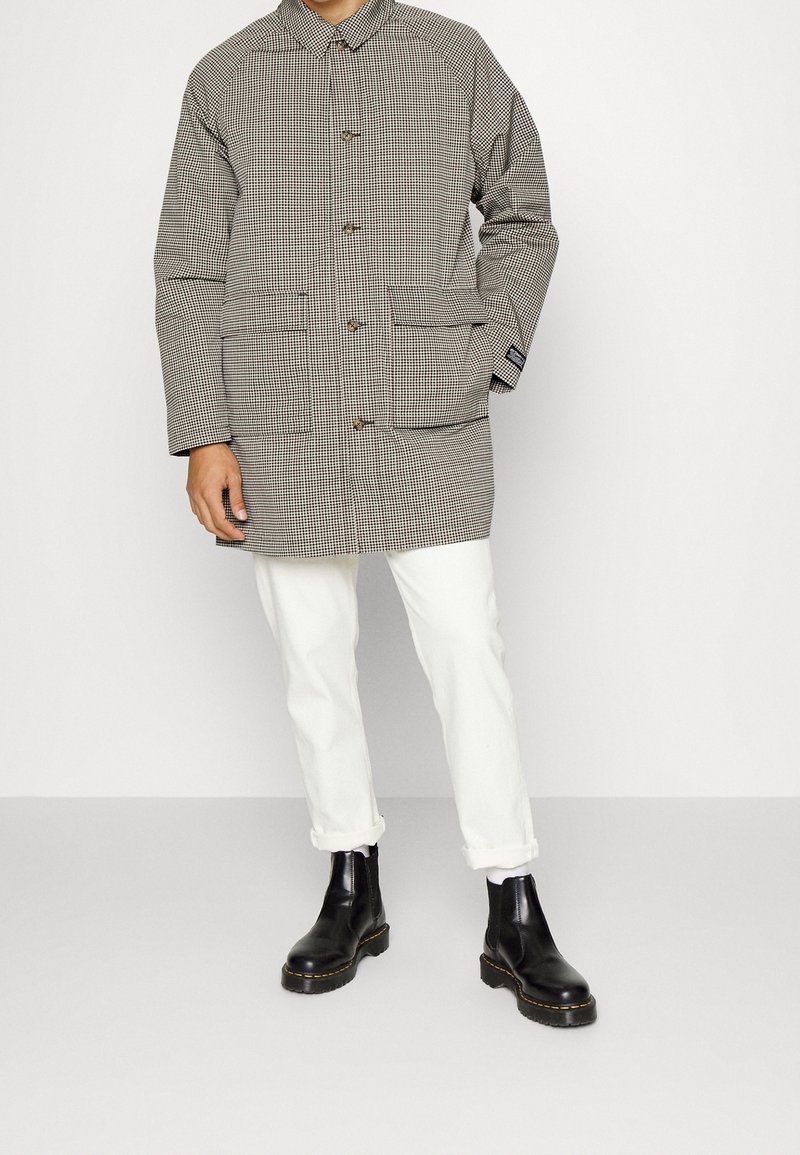 Abrigo de patrón a cuadros con dos bolsillos frontales, cierre de botones y cuello con solapas; combinado con pantalones blancos y botines negros.