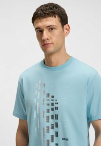 Camiseta de algodón azul claro con manga corta que presenta un estampado gráfico vertical en gris y negro. Cuello redondo y sutil logo de la marca en el dobladillo.