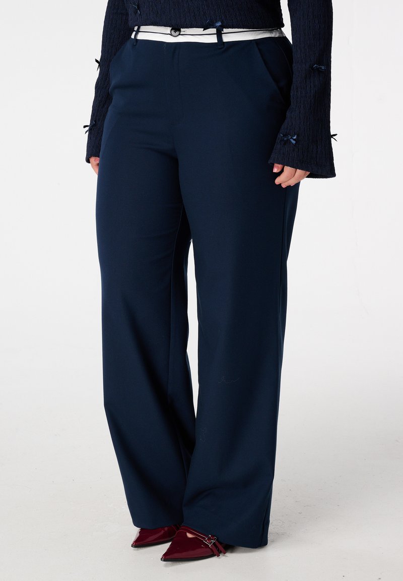 Marineblauwe wijde broek met een gladde textuur, voorzien van een witte riem en zijzakken. Gecombineerd met puntige bordeauxrode hakken.