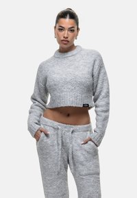 Vrouw met een bun in haar haar, gekleed in een lichtgrijze gebreide cropped trui en bijpassende joggingsbroek met strik, handen in de zakken, tegen een witte achtergrond.