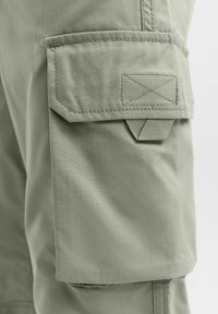 Detail vrecka na béžových nákladných nohaviciach s klopou zabezpečenou viditeľným prešitím a malým ťahákom pod klopou.