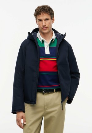Hombre con chaqueta azul marino sobre una camiseta de rugby de rayas verdes, rojas y azules, con pantalones beige y cinturón negro, de pie frente a un fondo blanco.