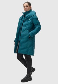 Teal puffer coat featuring a high collar, snap buttons, and side pockets. Worn with black jeans and chunky black boots. Clean, modern cut.