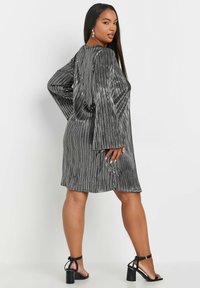 Skinande, silverfärgad klänning knälång med vertikala veck, vida ärmar och rund halsringning, i kombination med svarta klacksandaler.