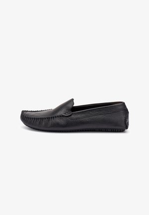 Mocasín negro de cuero sin cordones con costura estilo mocasín y suela texturizada, visto de lado sobre un fondo blanco.