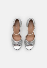 Silberne High Heel Sandalen mit glitzerndem Obermaterial, geschlossener Zehenpartie, verstellbarem Knöchelriemen und einem glatten, metallischen Absatz. Schlichte Designdetails vorhanden.