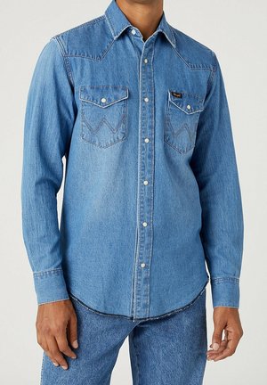 Mann trägt hellblaues Jeanshemd mit Knöpfen und aufgesetzten Brusttaschen sowie dunkelblaue Jeans, steht vor einfarbigem Hintergrund.