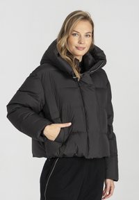 Czarna kurtka puchowa z wysokim kołnierzem i oversize’owym kapturem. Posiada pikowaną konstrukcję oraz boczne kieszenie. Materiał wydaje się błyszczący i izolowany.