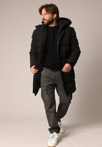 Zwarte pufferjas met capuchon, doorgestikt ontwerp en rits aan de voorkant. Draagt over een zwarte trui en grijze cargobroek, gecombineerd met witte sneakers.