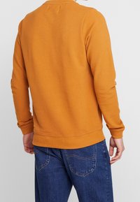 Sudadera naranja con un acabado texturizado, puños y dobladillo acanalados. Se combina visiblemente con jeans azules y un ajuste general relajado.