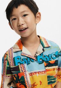 Spalvota trumpų rankovių marškinėlių palaidinė su spalvingų motyvų koliažu, įskaitant tropinius motyvus ir ryškų tekstą, su laisvu kirpimu ir apykakle.