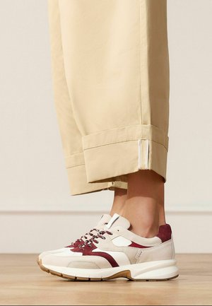 Beine in beigen kurzen Hosen, die beige und burgunderrote Wildledersneaker mit weißen Sohlen auf einem Holzboden tragen.