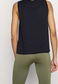 Zwarte mouwloze top met een textuurpatroon, voorzien van een hoge halslijn en een ontspannen pasvorm. Gecombineerd met olijfgroene aansluitende leggings.
