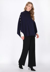 Poncho bleumarin cu guler ribat, accente cu butoni laterali și un design liber, fluid, asortat cu pantaloni largi negri și cizme cu toc.