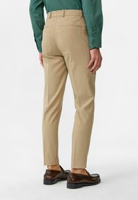 Beige broek gemaakt van een gladde stof, met een aansluitend ontwerp en achterzakken. Gekleed met donkerbruine schoenen, staand tegen een neutrale achtergrond.