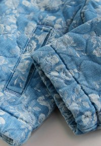 Světlomodrá džínová látka s bílým květinovým vzorem, s výraznou texturou a prošíváním. Látka je složena tak, aby byl vzor dobře viditelný.