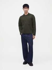 Jeune homme debout portant une chemise boutonnée vert foncé avec deux poches poitrine, un jean bleu large et des chaussures noires sur fond blanc.