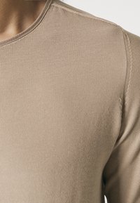 Chemise beige à manches longues avec un tissu texturé côtelé et un col rond. Le matériau semble doux et légèrement extensible.