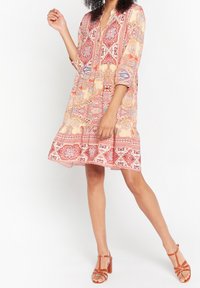 Långärmad klännning i knälängd i ljusrosa med invecklade blommönster och geometriska mönster. Lättviktig tygkvalitet, plisserad fåll, bärs med bruna klacksandaler.