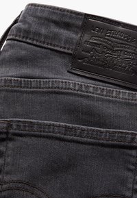 Vaqueros de denim gris oscuro con una superficie texturizada, presentando una etiqueta de cuero negra y detalles de costura visibles en el bolsillo trasero y la cintura.
